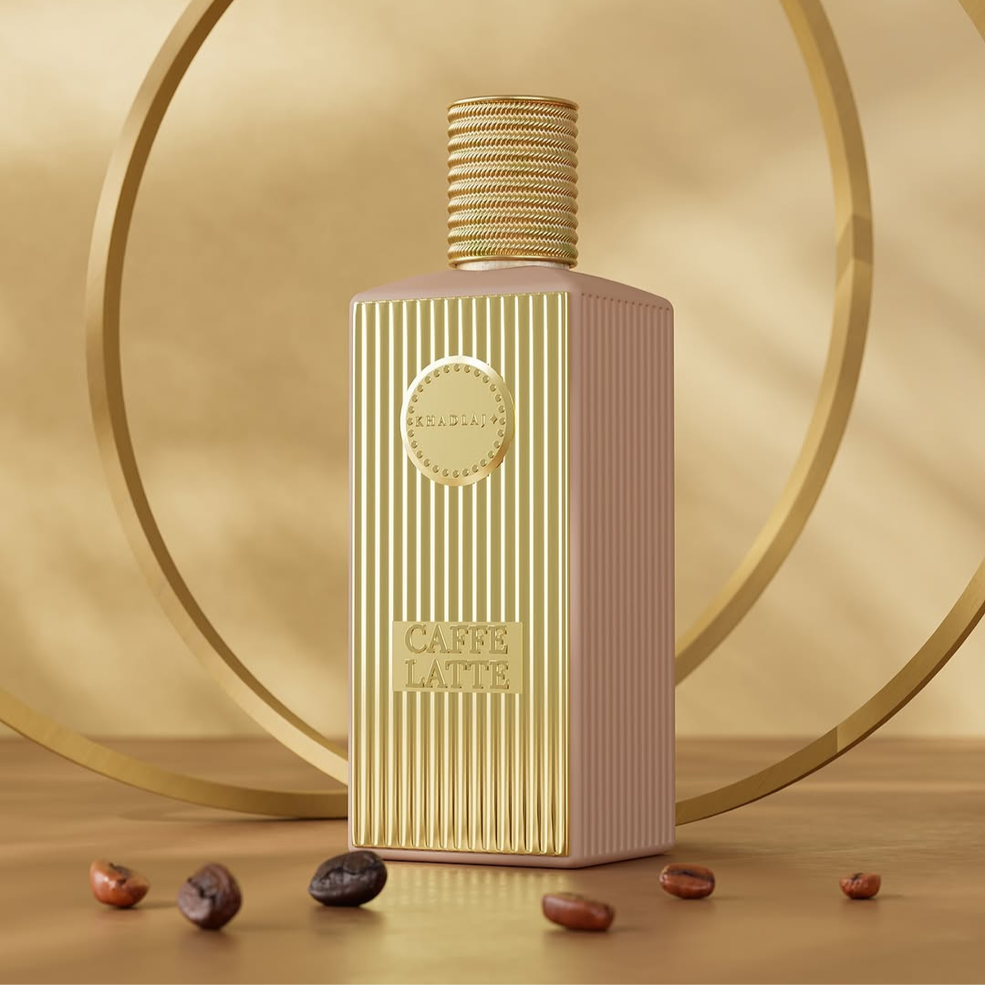 A rectangular Khadlaj Caffe Latte 100ml EDP bottle with vertical grooves sits on a reflective surface, exuding a coffee fragrance surrounded by scattered coffee beans and two large intersecting gold rings in the background.