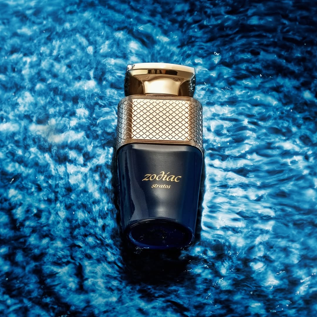 A dark blue Zodiac Stratos 100ml EDP bottle by Paris Corner, featuring a textured gold cap, sits against a vibrant swirling water backdrop, capturing earthy notes that embody celestial and terrestrial realms.