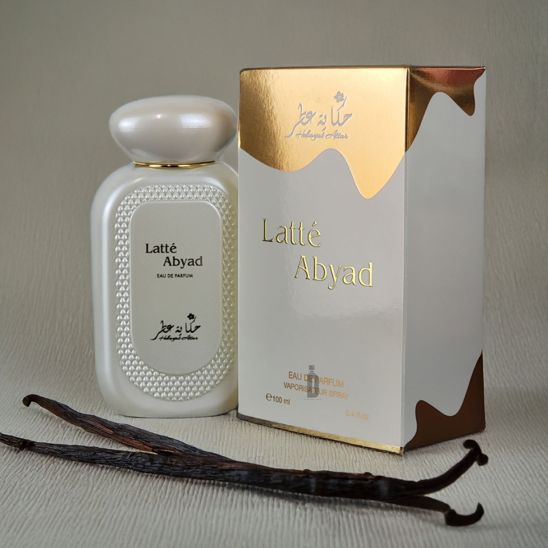 A bottle of Latte Abyad 100ml EDP by Hekayat Attar sits next to its elegant box on a textured surface. The white and gold packaging with decorative pearls exudes luxury, while vanilla pods underscore the indulgent fragrance notes.