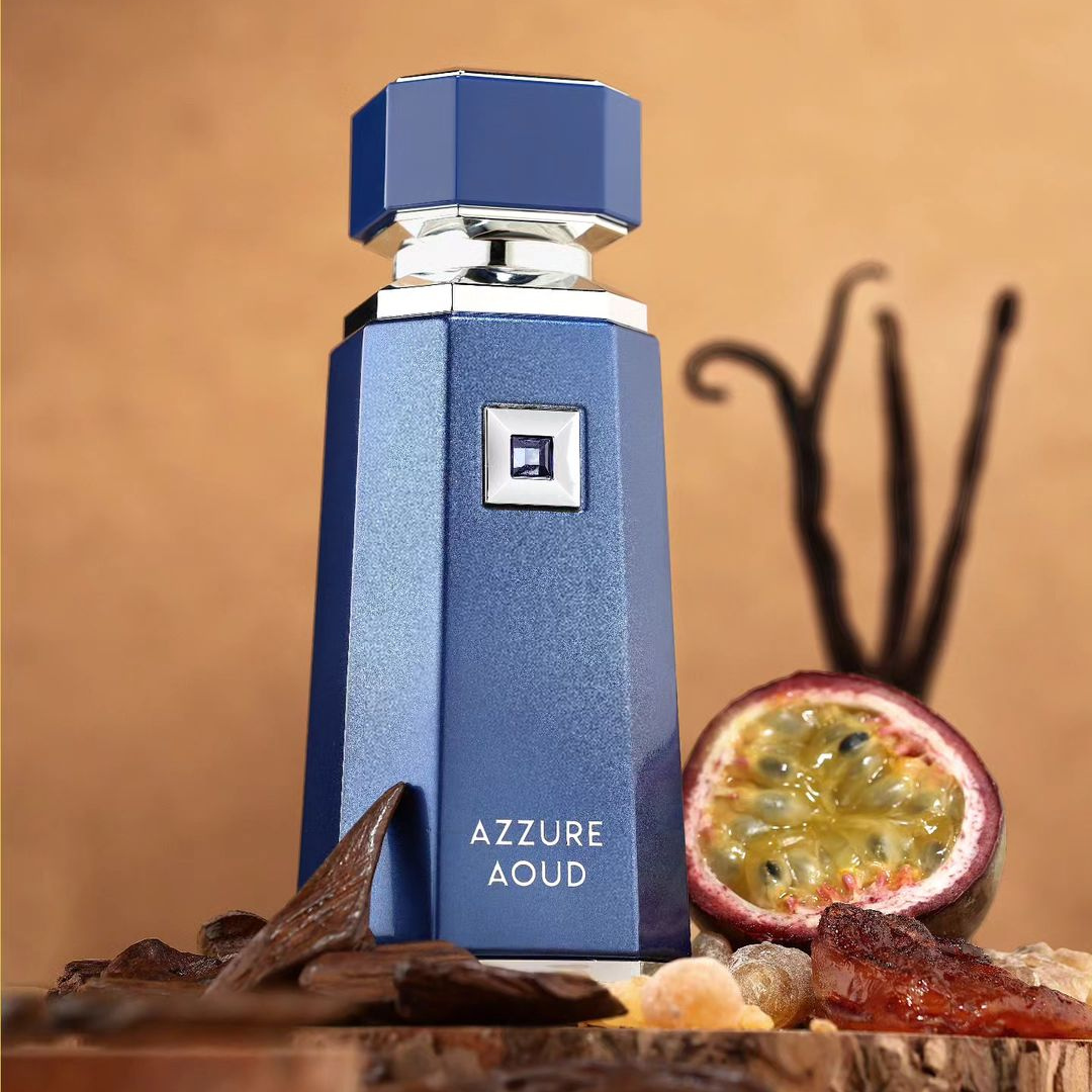 A blue, geometric bottle of Azzure Aoud 100ml EDP from Fragrance World is displayed on a wooden surface. This oud perfume is accompanied by aromatic components like a halved passion fruit, vanilla beans, and pieces of resin, crafting an exotic fragrance set against a warm beige background.