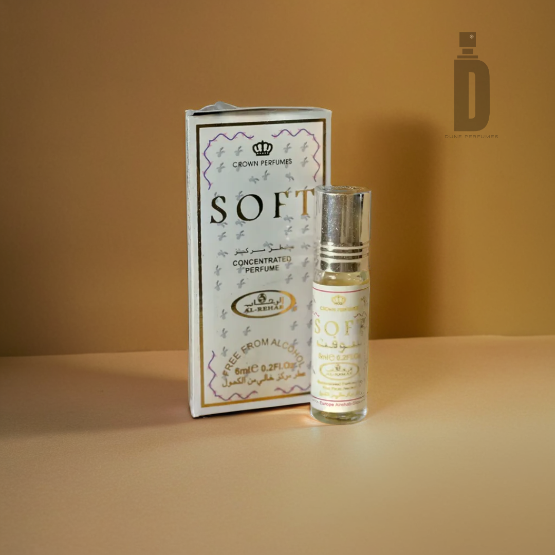 A bottle of "Soft - Al Rehab" concentrated perfume by Al Rehab rests beside its box on a beige surface, emitting a gentle aroma. The packaging showcases an elegant design with cursive text and a small crown emblem at the top, suggesting luxurious vanilla notes. The backdrop is in a simple, warm tone.