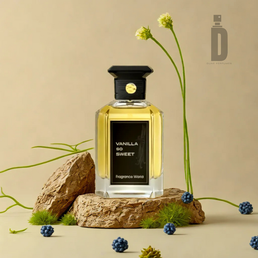 A bottle of Fragrance World's "Vanilla So Sweet" 100ml EDP, topped with a black cap, is elegantly displayed on a stone surface amidst small blue berries, grass, and delicate flowers against a beige backdrop. The fragrance notes offer a sweet scent that perfectly complements its captivating setting.