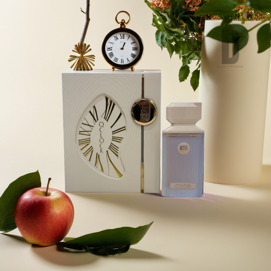 An elegant display featuring the O'clock 100ml EDP by Ahmed Al Maghribi includes a pocket watch, an apple, lush green leaves, and a vase of flowers against a neutral backdrop. Experience the fragrance that encapsulates the excitement of new adventures with its captivating citrus burst.