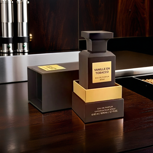 A bottle of "Vanille En Tobacco 80ml EDP - Fragrance World" sits partially out of its packaging on a dark wooden surface, releasing a blend of vanilla and tobacco scents. The backdrop features a sleek metallic element from the kitchen setting.