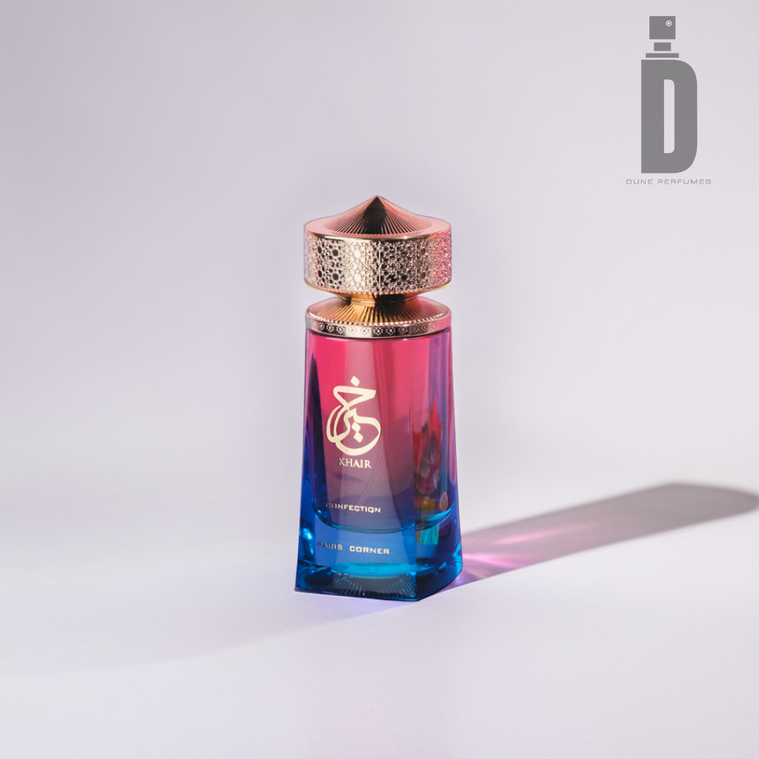 A perfume bottle with an ombre design, transitioning from blue at the base to pink at the top, features a gold cap with intricate detail. The bottle has gold Arabic script and English inscription "Khair Confection" and "AL HARAMAIN." A "D" logo on the top right says "Dune Perfumes," exuding floral elegance. The product name is Khair Confection 100ml EDP by Paris Corner.
