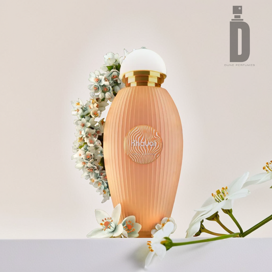 A peach-colored perfume bottle featuring a ribbed texture and gold accents, labeled Khayal 100ml EDP by Paris Corner, is set among small white flowers, releasing a bergamot fragrance. The bottle boasts a spherical cap and floral wreath design that suggest a woody warmth. The Paris Corner logo is prominently displayed in the upper right corner.