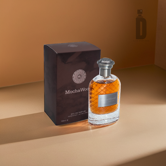 A glass bottle of Mocha Wood 100ml EDP by Fragrance World, filled with amber liquid, rests next to a dark brown box featuring the brand name in white. Both are tastefully placed on a tan surface, where soft shadows enhance the fragrance's elegant charm.