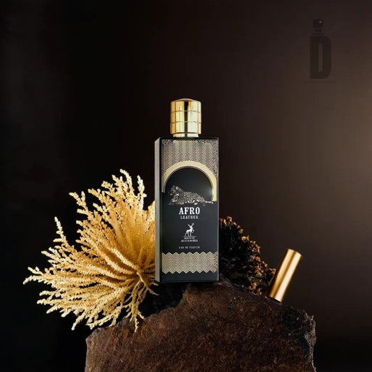 An 80ml bottle of Afro Leather Eau de Parfum by Maison Alhambra, featuring a black and gold design, rests majestically on a rock. The background showcases dark tones accented by light-colored, coral-like branches encircling the bottle. This bold fragrance includes a striking gold cap and intricate gold detailing, enhancing its exotic appeal.