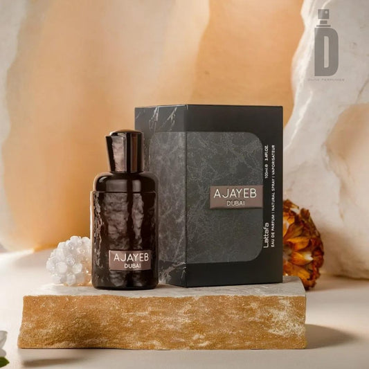 A bottle of Ajayeb Dubai 100ml EDP by Lattafa stands next to its matching black and gray box on a stone slab. The background features a white, abstract texture with a low arrangement of flowers and white crystals, epitomizing the opulence and mystery of this luxury fragrance. The Lattafa logo is visible at the top right corner.