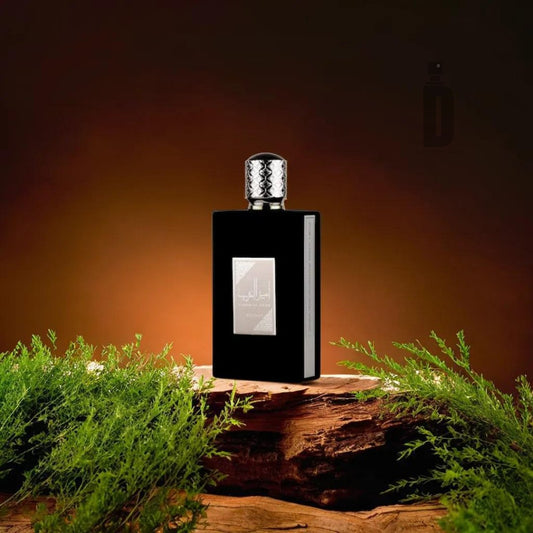 A sleek black perfume bottle with a textured silver cap, embodying regal sophistication, rests on a wooden surface surrounded by green foliage. Set against a warm, gradient brown background, the light subtly highlights the elegant design and reflective label of this Ameer al Arab 100ml EDP by Lattafa.