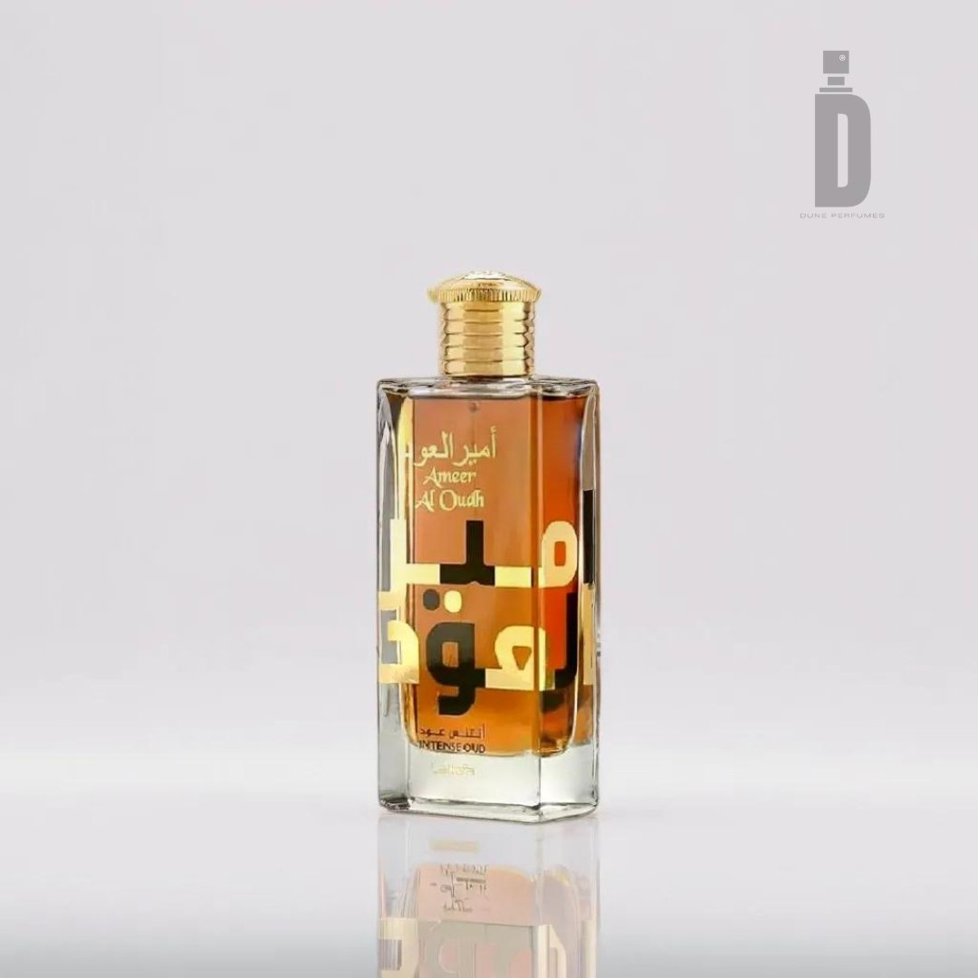 A clear rectangular perfume bottle with gold accents, featuring the text "Ameer Al Oud Intense 100ml EDP" on the front in Arabic and English. The bottle is filled with a dark amber liquid embodying a rich oud fragrance and has a gold cap. The brand name "Lattafa" is visible at the top right corner.