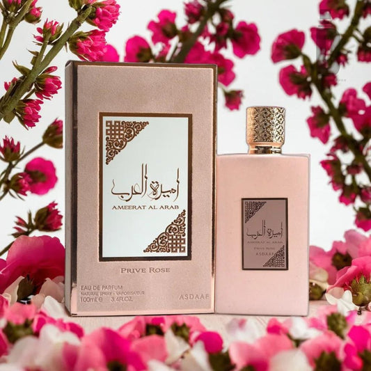 A perfume bottle and its box labeled "Ameerat Al Arab Prive Rose 100ml EDP" by Lattafa, exuding a luxurious fragrance, are surrounded by blooming pink flowers. The box and bottle feature an elegant design with Arabic text adorned with decorative patterns, capturing the essence of a floral bouquet.