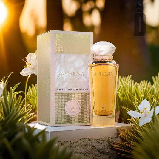 A bottle of Athena 100ml EDP - Maison Alhambra sits elegantly next to its matching light green and white packaging box. The scene is set outdoors in soft, warm lighting, surrounded by green foliage and white flowers, creating a serene and luxurious ambiance that highlights this divine fragrance from Maison Alhambra.