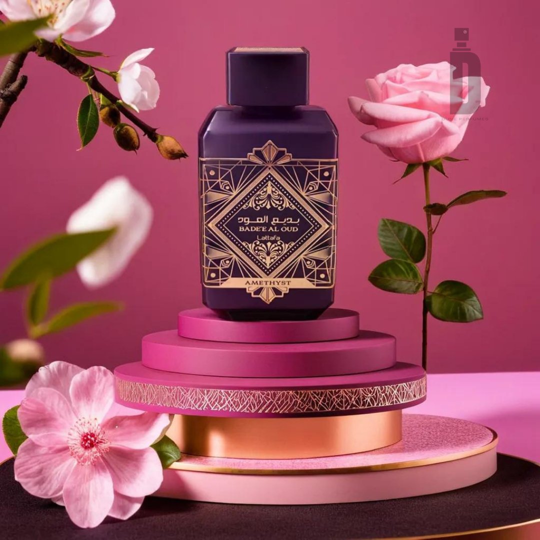 A bottle of Badee Al Oud Amethyst 100ml EDP - Lattafa stands on a multi-tiered pedestal with pink and gold accents. The background features a gradient pink backdrop and blooming flowers, adding elegance to the display. The Turkish rose notes intertwined with agarwood (oud) create an enchanting Amethyst fragrance by Lattafa.
