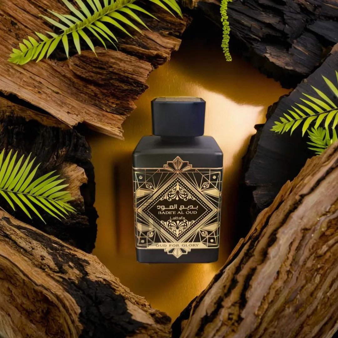 A perfume bottle labeled "Badee Al Oud (Oud For Glory) 100ml EDP - Lattafa" is positioned at the center of the image, surrounded by pieces of agarwood and green fern leaves. The bottle, exuding the essence of a luxury fragrance with its intricate geometric design and dark, luxurious appearance, is set against a golden background.