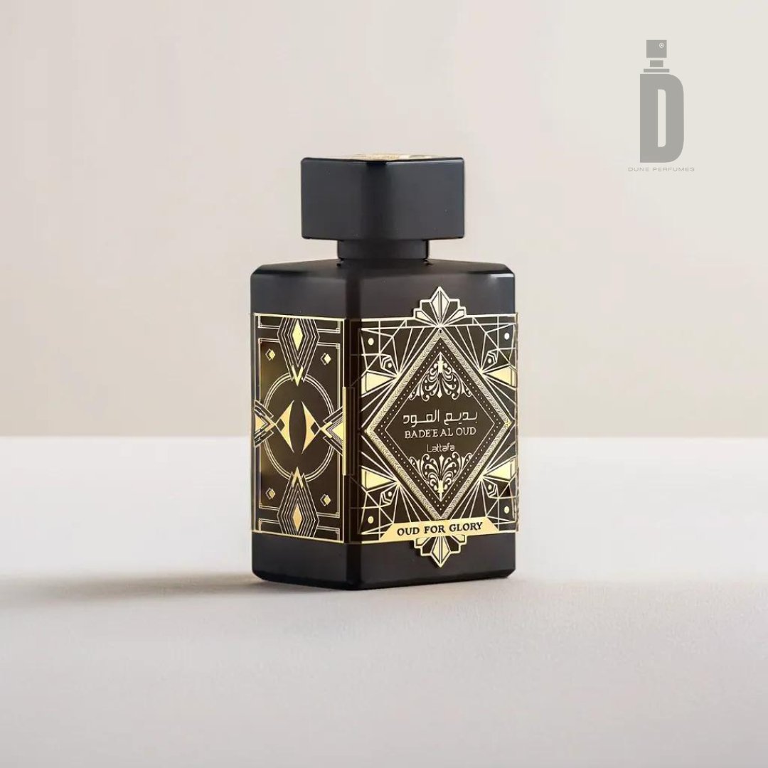 A black rectangular perfume bottle with intricate gold patterns and the text "Badee Al Oud" and "Oud For Glory" on the label. This luxury fragrance, named Badee Al Oud (Oud For Glory) 100ml EDP by Lattafa, features a gold-trimmed cap and sits on a gray surface against a beige background. The Lattafa logo is in the top right corner of the image.