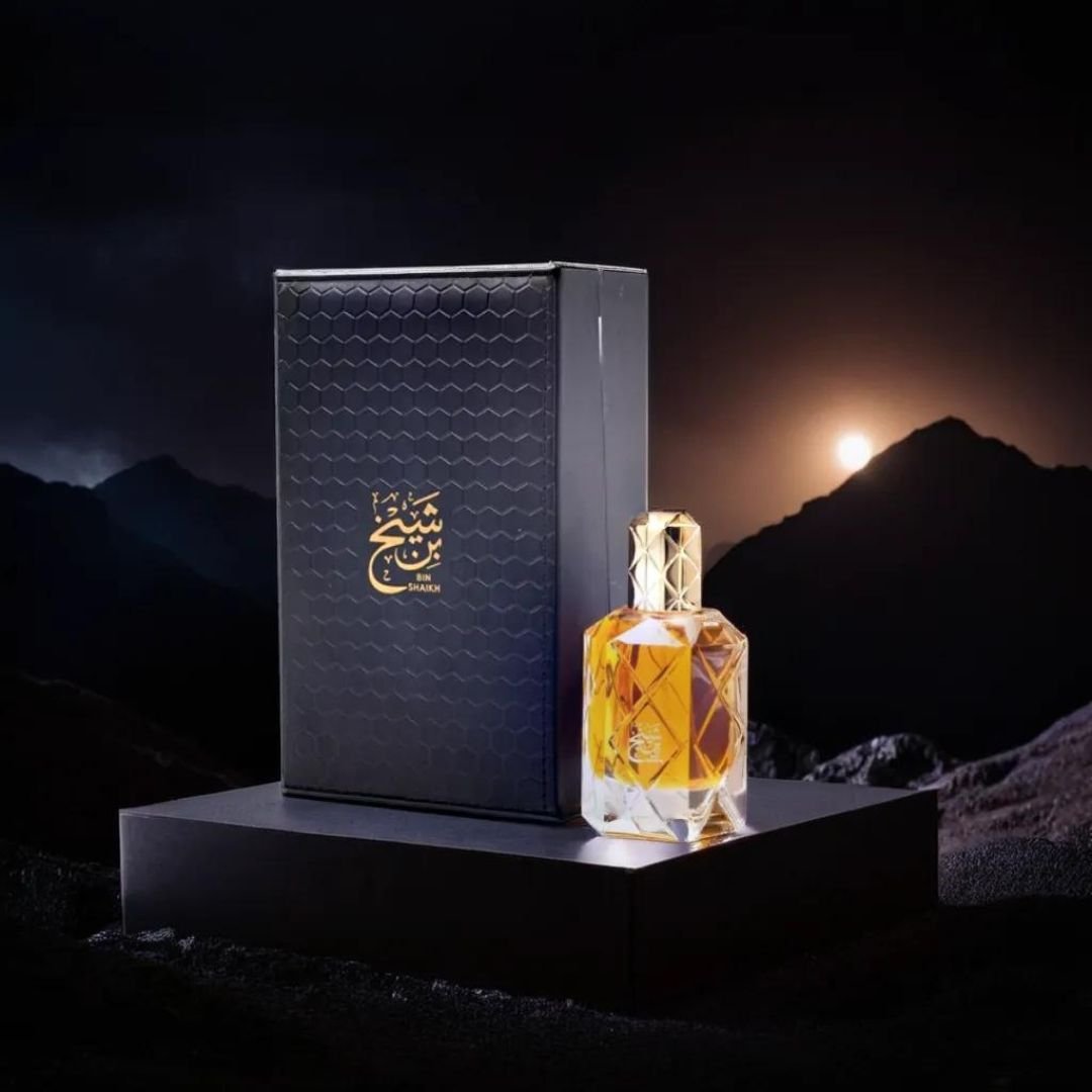 A 90ml perfume bottle from Ahmed Al Maghribi, named Bin Shaikh, features a diamond-patterned design and golden cap, resting beside its sleek black box adorned with elegant golden Arabic script that embodies Arabic grandeur. The background showcases a dramatic, dark mountainous landscape illuminated by a setting sun. This is Bin Shaikh Eau de Parfum—a truly luxurious scent.