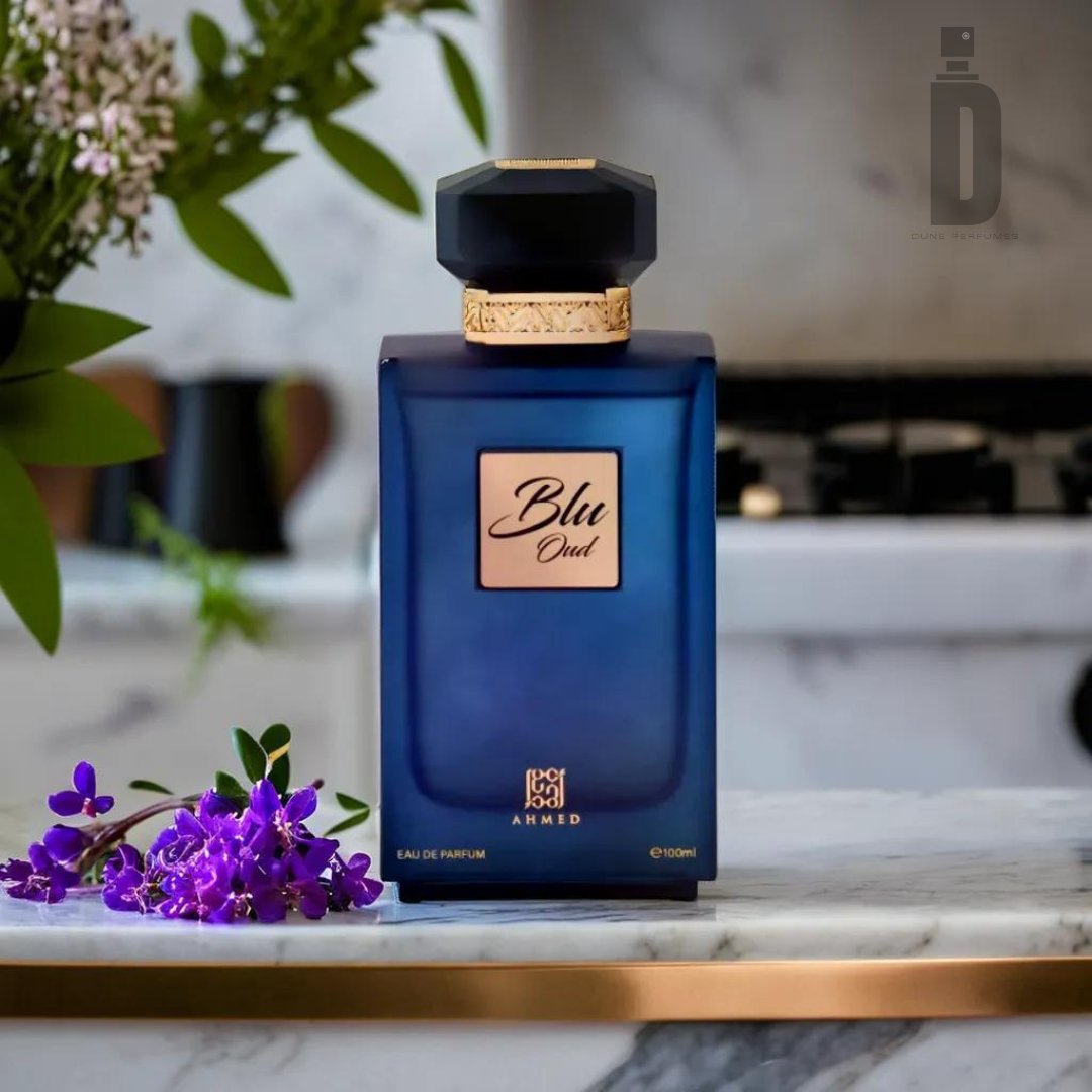 A bottle of Blu Oud 100ml EDP by Ahmed Al Maghribi, featuring a blue exterior with a black cap and gold detailing, is elegantly placed on a marble surface. The sophisticated fragrance is complemented by a small bouquet of purple flowers beside it. In the background, blurred elements of a kitchen with plants and an appliance add depth to the scene.