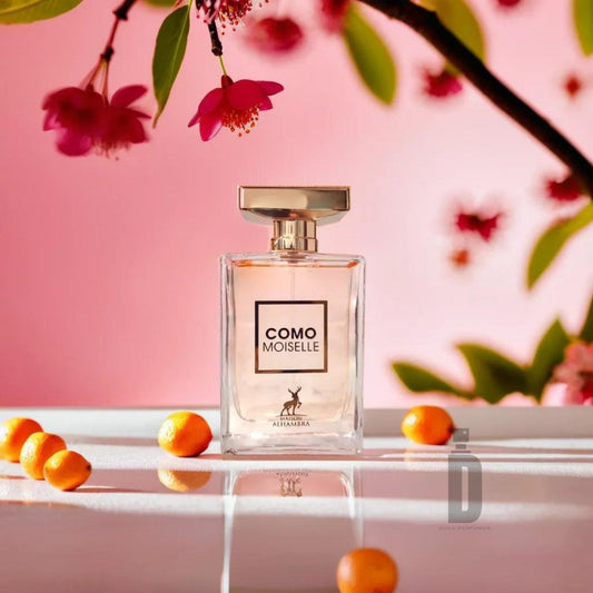 A perfume bottle labeled "Como Moiselle 100ml EDP" by Maison Alhambra rests on a white surface, surrounded by small orange fruits. Pink flowers on slender branches hang nearby, with a soft pink background setting a delicate and elegant ambiance that perfectly complements its feminine fragrance and floral notes.