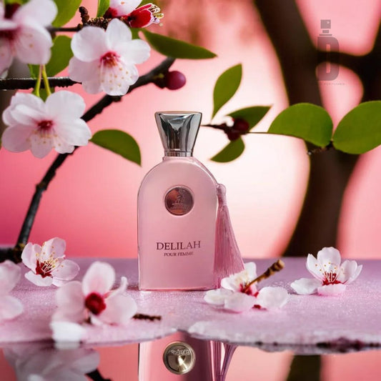 A pink bottle of Delilah 100ml EDP - Maison Alhambra stands on a reflective surface surrounded by delicate cherry blossoms. The background is a gradient of pink hues, enhancing the elegant and feminine presentation of the Turkish rose and white musk-infused perfume from Maison Al Hambra.