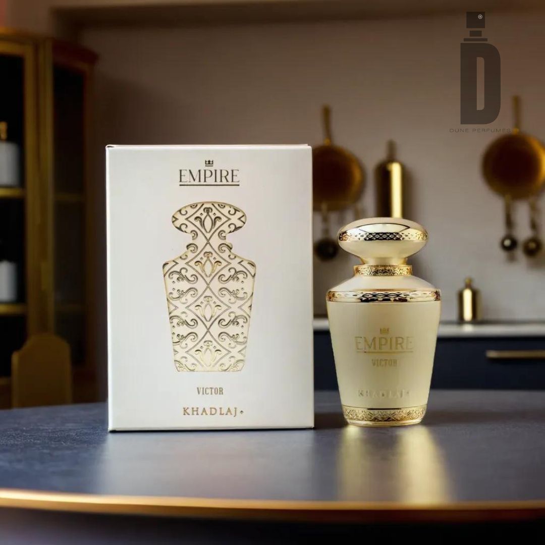 A perfume bottle labeled "Empire Victor 100ml EDP - Khadlaj" sits on a dark surface in front of a matching box adorned with an ornate gold pattern. The background reveals a modern kitchen with hanging utensils and minimalist decor, perfectly capturing the essence of this luxurious fragrance by Khadlaj.