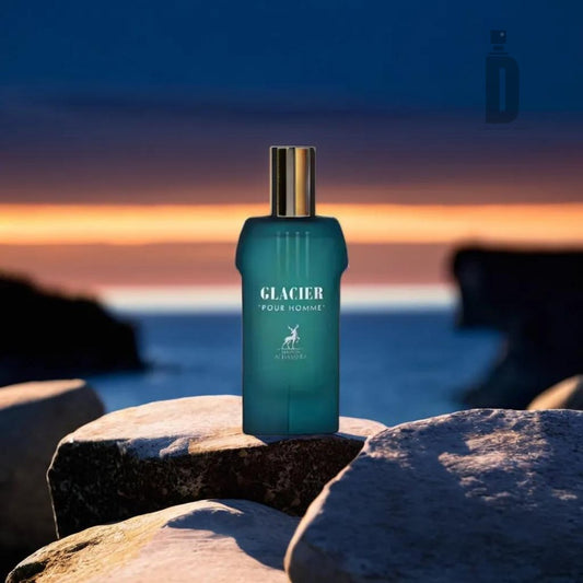 A teal bottle of Glacier Pour Homme 100ml EDP by Maison Alhambra stands on a rock with a golden cap, set against a backdrop of a serene dusk sky over the ocean, framed by silhouette cliffs. The image emphasizes the Maison Al Hambra men's fragrance's connection with nature and freshness.