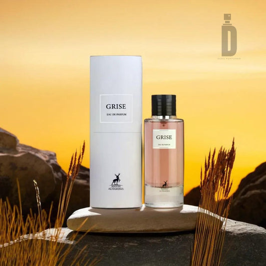 A bottle of Grise 100ml EDP - Maison Alhambra, embodying timeless sophistication, is placed on a wooden platform. Its cylindrical white packaging sits elegantly beside it. The background features a rocky landscape with tall grass against a warm, golden sky, hinting at its floral woody notes.