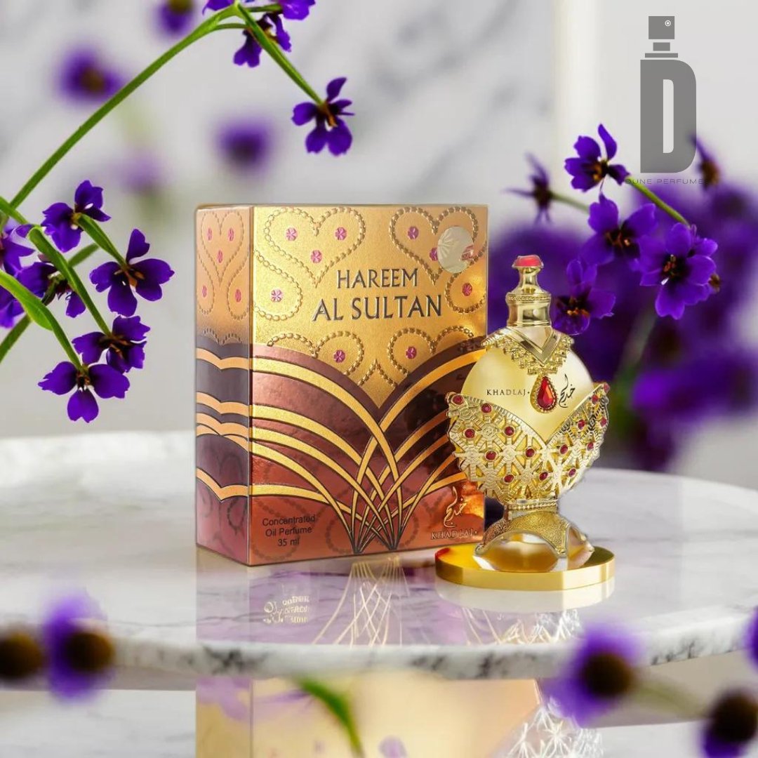 A decorative perfume bottle labeled "Hareem Al Sultan Gold 35ml Perfume Oil - Khadlaj" sits on a marble surface with a matching ornate box behind it. The gold and red design of the bottle and box is intricate and detailed. Purple flowers are blurred in the background, adding a touch of elegance to this non-alcoholic formulation by Khadlaj.