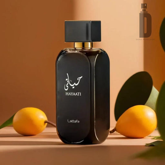 A perfume bottle labeled "Hayaati 100ml EDP - Lattafa" by Lattafa is placed on a surface with a soft focus background. The sleek, black bottle with a square cap exudes sophistication. Two yellow-orange fruits and green leaves surround it, adding a touch of natural vibrancy to the fragrance's presentation.