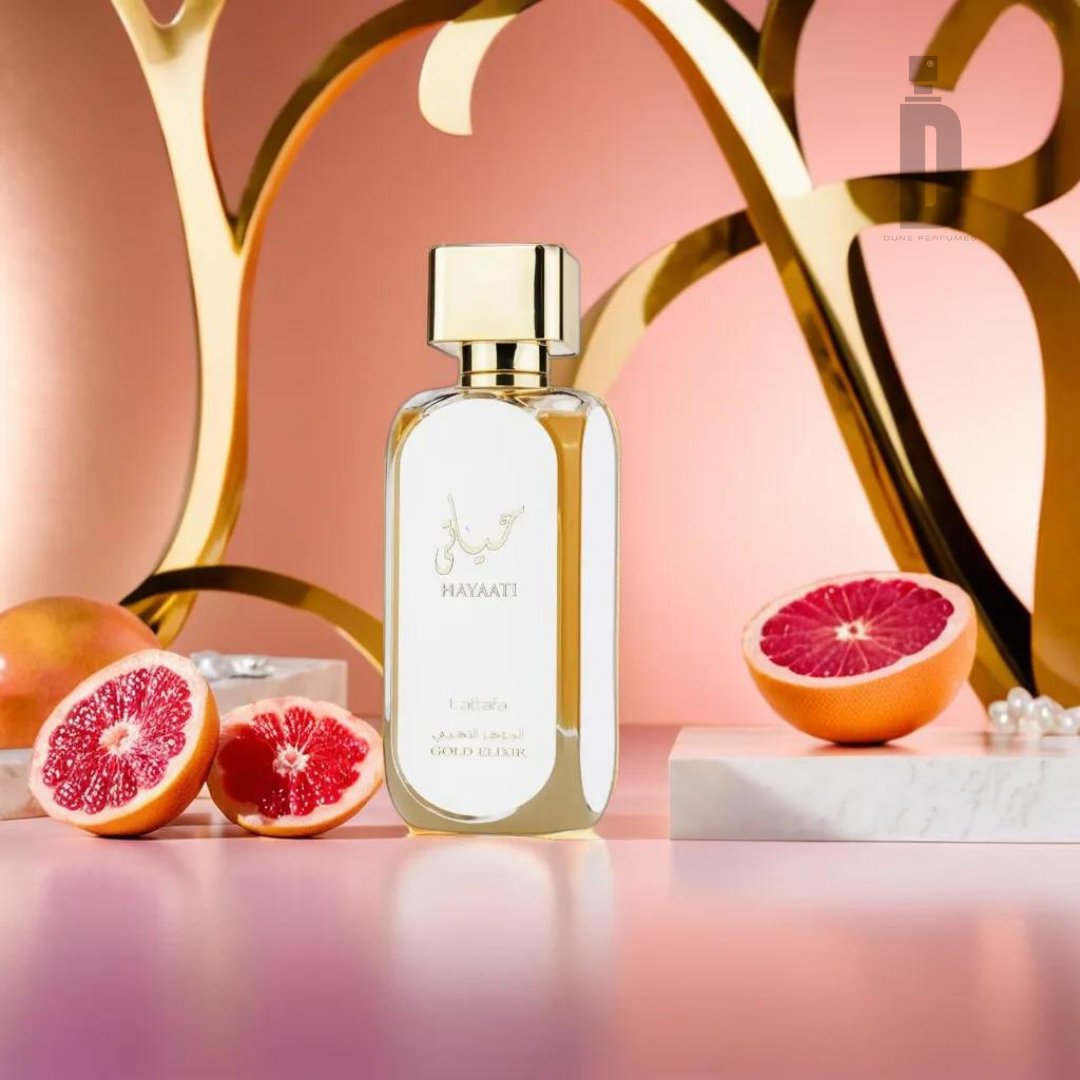 A bottle of Lattafa's Hayaati Gold Elixir 100ml EDP, celebrated for its luxurious fragrance, is showcased against a pink and peach-toned background. The bottle features a gold cap and label. Beside it are two halved grapefruits, with one resting on a marble slab and the other placed directly on the surface. The backdrop is enhanced with abstract golden shapes.