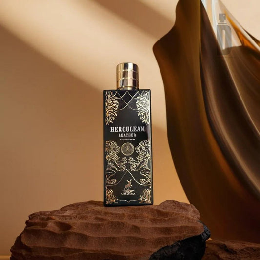 A sleek, rectangular bottle of Herculean Leather 80ml EDP by Maison Alhambra stands on a textured rock. The fragrance features ornate, gold detailing with mythical illustrations. A flowing, brown fabric swooshes in the background, adding a touch of strength and elegance.