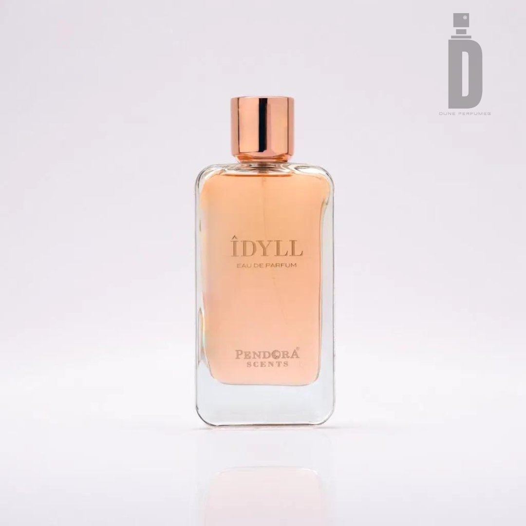 A clear rectangular bottle of Idyll 100ml EDP by Paris Corner, featuring a rose gold cap and peach-colored liquid. The sophisticated scent and aromatic layers are embodied in its design. The product logo and brand name are printed in gold on the front, against a plain white background.