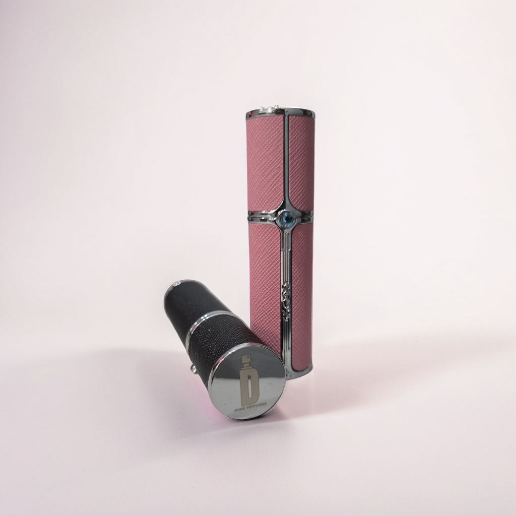 Two 5ml Travel Spray bottles from Dune Perfumes, each with a textured exterior, are positioned against a light pink background. One bottle is upright and covered in pink, while the other lies horizontally with a black exterior and a reflective silver cap.