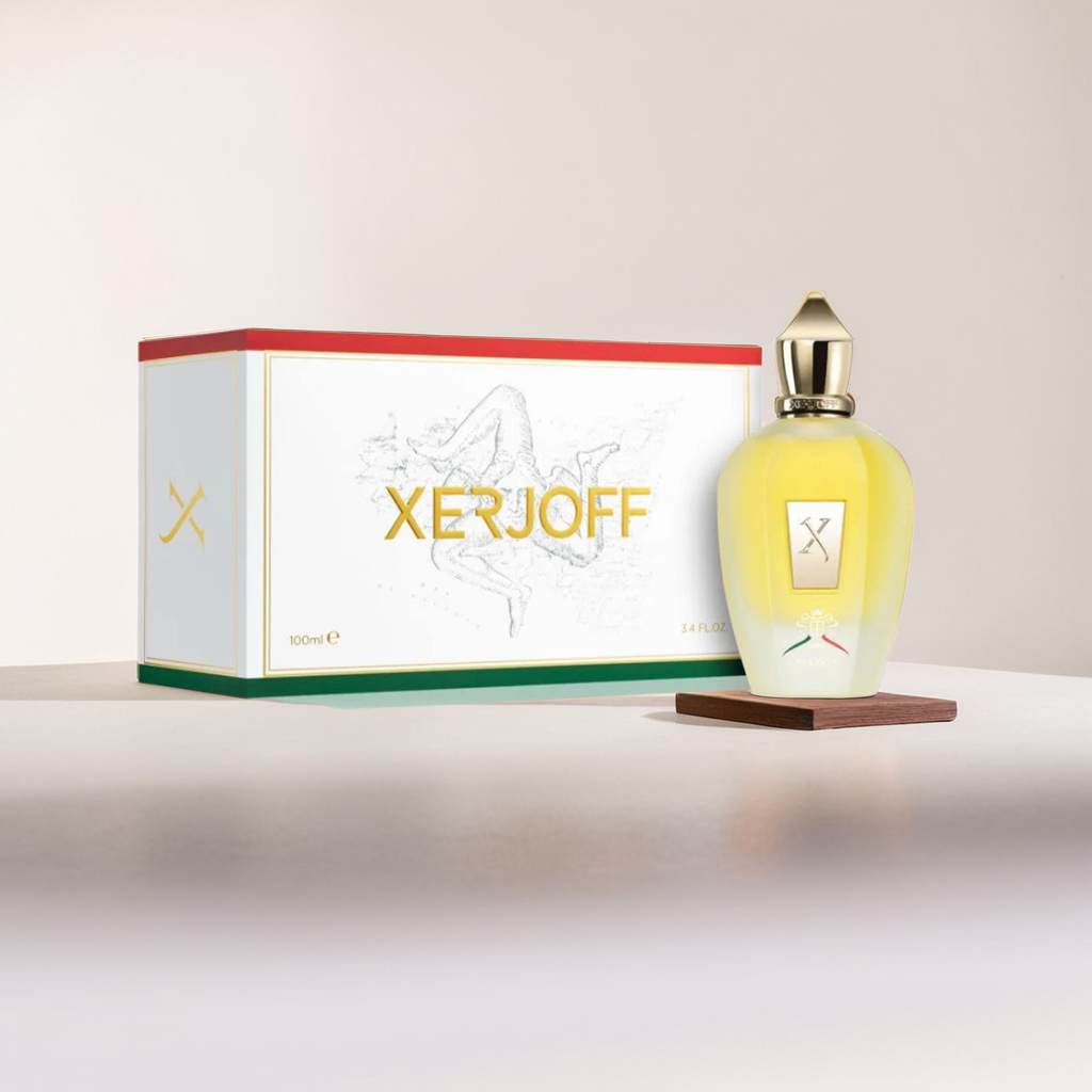 A Naxos XJ 1861 100ml EDP bottle by Xerjoff, with a gold top and yellow liquid, elegantly stands beside its box. The box displays the brand name in gold and an angel design on a soft beige background, promising an exquisite scent experience.