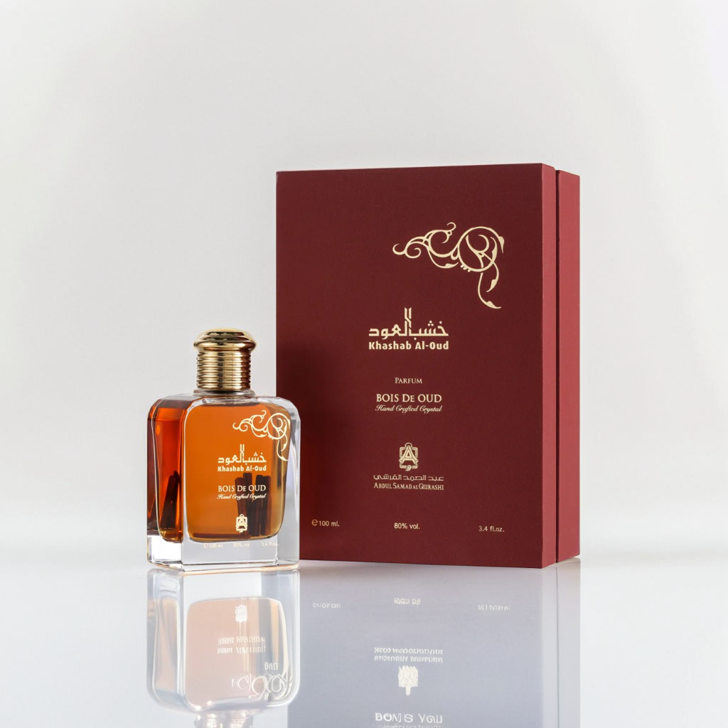 A Khashab Al-Oud 100ml EDP by Abdul Samad Al Qurashi, encased in a glass bottle with a golden cap, is positioned before a maroon box showcasing Arabic script and gold designs. The luxurious packaging highlights the agarwood scent against a plain white backdrop.