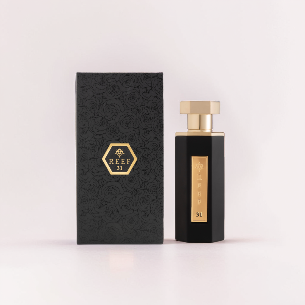 A sleek black perfume bottle with a gold cap and a label reading "Reef (31)" stands beside a matching black box with rose patterns. A crown graphic hints at saffron and amber notes.