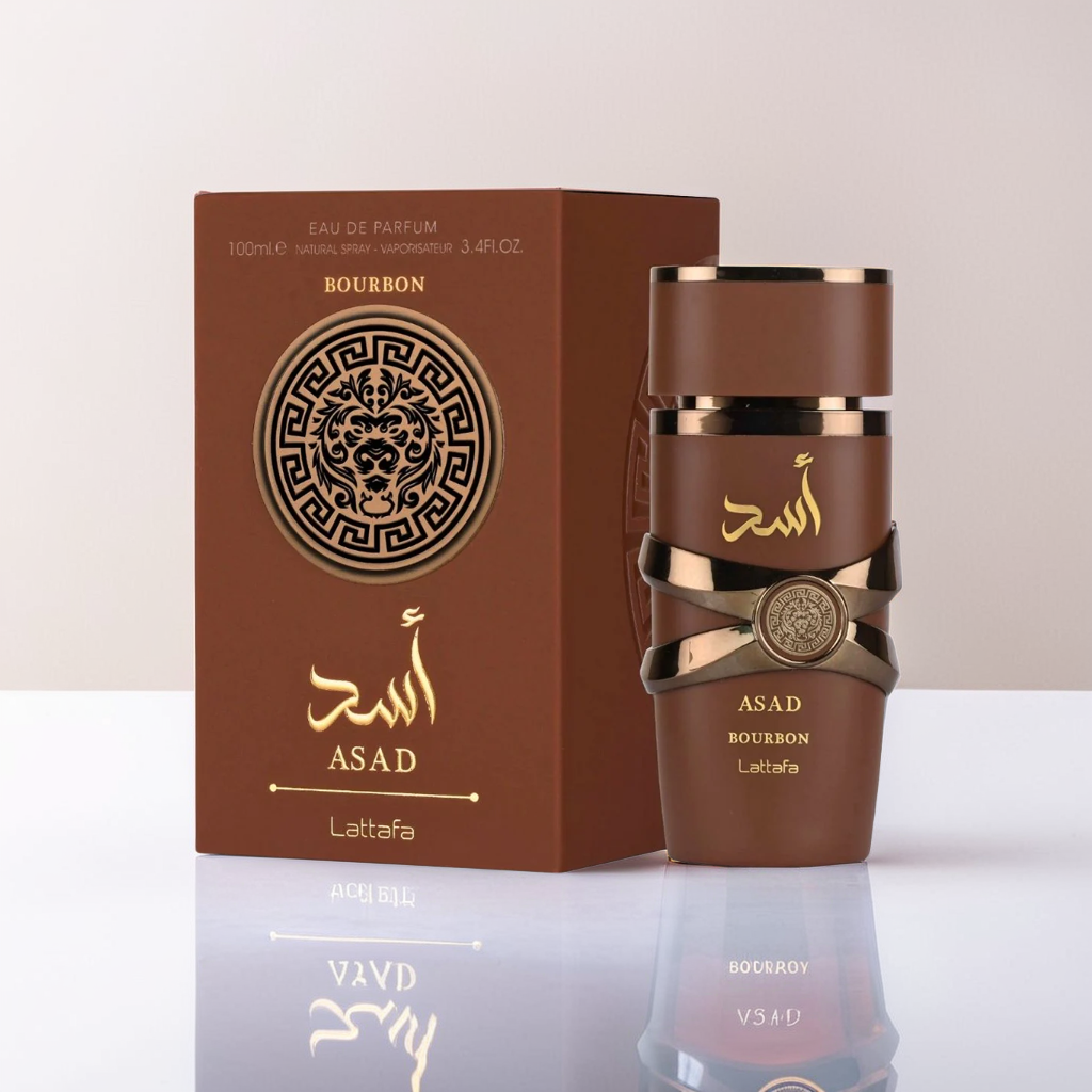 A brown perfume box labeled "Asad" and adorned with golden Arabic script stands beside an elegant bottle showcasing a metallic cross design. The label reads "Asad Bourbon 100ml EDP - Lattafa." The background features a simple gray surface.