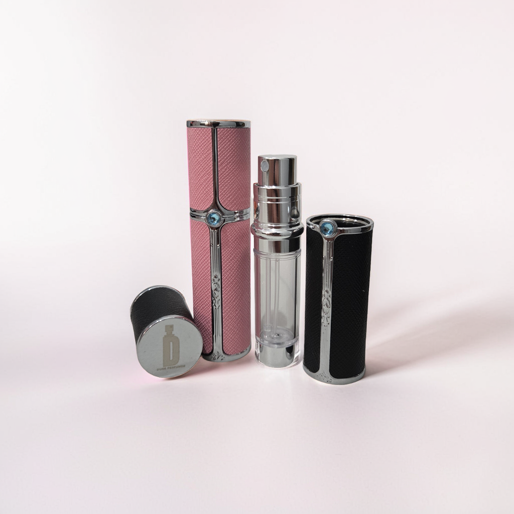 Two elegant 5ml Travel Sprays by Dune Perfumes are displayed against a soft pink background. One is pink and the other black, both featuring metallic details and a striking blue gemstone accent. The black spray is open, showcasing the clear glass bottle inside.