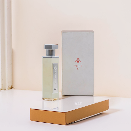 The "Reef 33 White 100ml EDP - Reef" perfume and its matching box, both featuring a simple modern design with a geometric logo and oud hints, elegantly rest on a white surface against a soft background.