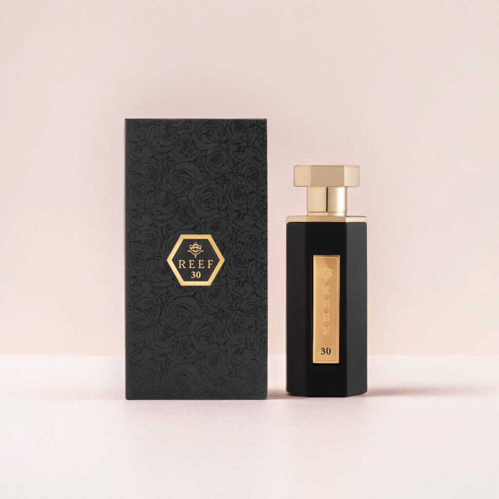 A sleek black Reef (30) 100ml EDP bottle with a gold label is beside a floral-patterned black box, both adorned with a gold hexagonal logo. They embody the elegance of Reef's refined fragrance against a soft, pale pink background.