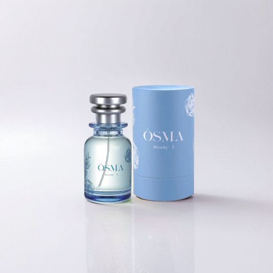 A glass bottle of Osma Woody S 75ml EDP with a metallic cap rests beside a cylindrical light blue box on a reflective white surface. The packaging features subtle floral graphics, suggesting the earthy woods that characterize its scent.