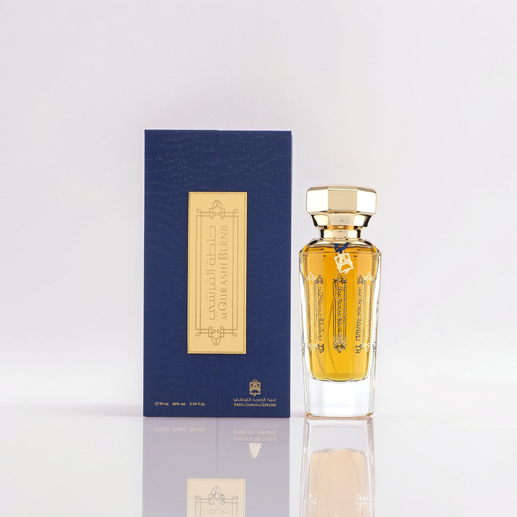 The Al Qurashi Blend 90ml EDP by Abdul Samad Al Qurashi features a glass bottle with amber liquid and a gold cap, set beside a blue box embellished with gold text and designs. Reflecting on the white surface, the presentation suggests luxury and hints at its oud-infused fragrance.