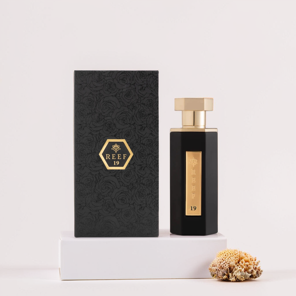 The Reef (19) 100ml EDP features a sleek black bottle with a gold cap and label beside a matching textured floral pattern box. The "REEF 19" logo is in gold. A nearby coral on a white surface hints at its delicate white musk floral scent.