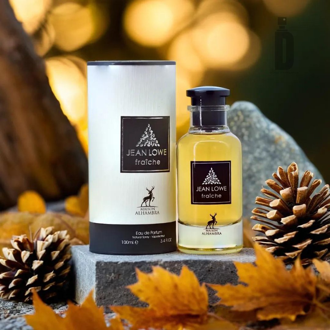 A bottle of Jean Lowe Fraiche (Nouveau) 100ml EDP - Maison Alhambra and its packaging sit surrounded by pinecones and autumn leaves. The fragrance bottle is clear with a dark cap, while the cylindrical packaging is white featuring the same branding. The scene evokes a captivating allure and timeless elegance, reminiscent of a natural, autumnal ambiance.