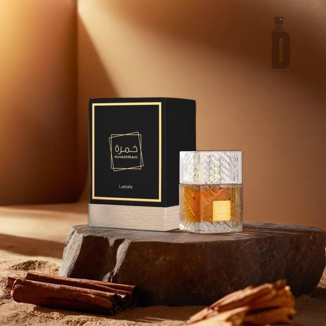 A stylish bottle of Khamrah 100ml EDP by Lattafa, filled with amber liquid, sits on a wooden slab. The black packaging box with a gold label stands behind it. This setup is arranged on a sandy surface adorned with cinnamon sticks and sweet spices, all bathed in warm light. Luxurious base notes elevate the fragrance of Khamrah.