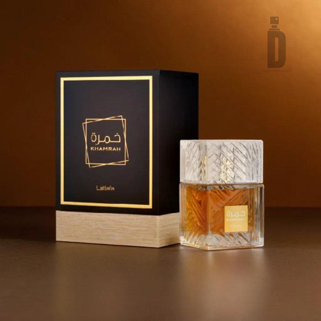 An image of the "Khamrah 100ml EDP - Lattafa" fragrance bottle and its packaging. The clear bottle contains amber liquid and features an intricate, textured design. The black and gold box is labeled "KHAMRAH" and "Lattafa," placed beside the bottle against a bronze background, suggesting luxurious base notes with sweet spices.