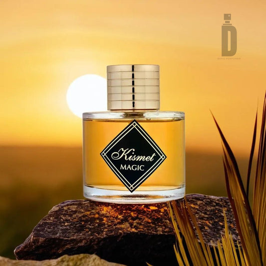 A glass bottle of Kismet Magic (Angel) 100ml EDP - Maison Alhambra with a gold cap sits on a rock. The background shows a serene sunset over a blurred landscape, with the sun casting a warm, golden glow. An enchanting aroma fills the air as the Maison Al Hambra logo shines in the top-right corner, epitomizing luxury fragrance.