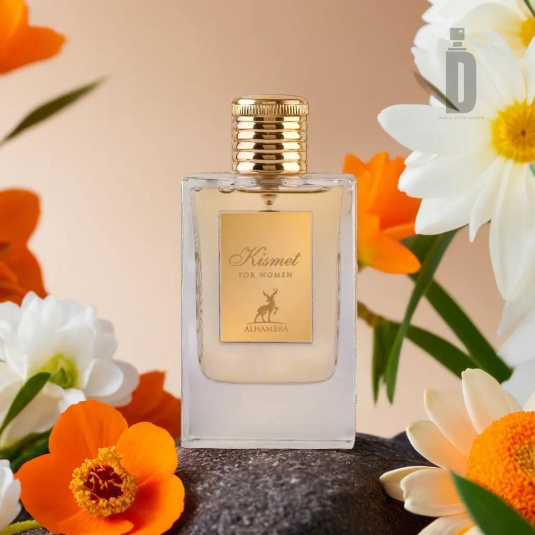 A perfume bottle labeled "Kismet Women 100ml EDP - Maison Alhambra" by Maison Al Hambra is placed on a rock adorned with orange and white flowers. The bottle boasts a sleek design with a golden cap and label, set against a warm gradient background. A faint atomizer logo is visible in the top right corner, promising an enchanting aroma.