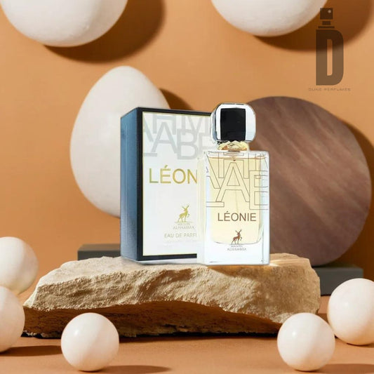 A bottle of Leonie (Libbra) 100ml Eau de Parfum by Maison Alhambra is positioned on a textured stone with its matching box in the background. The setup includes other decorative elements like spheres and geometric shapes, set against a warm, brown backdrop. The air hints at floral fragrances and warm vanilla.