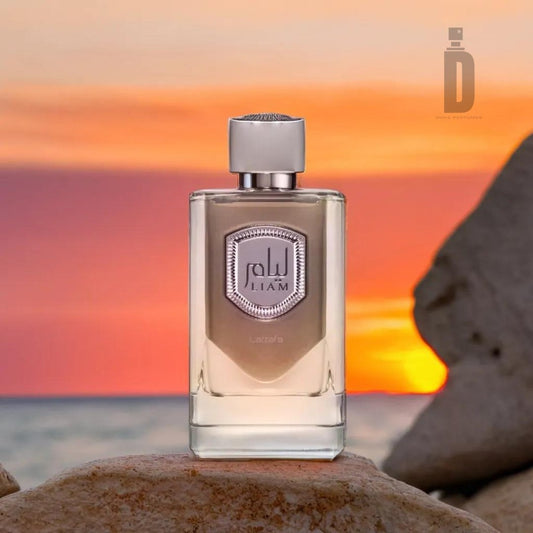 A clear perfume bottle with a silver cap and a hexagonal label that reads "JILAM" stands on a rocky surface. The background showcases a serene sunset over the ocean, with warm hues of orange and pink, and a silhouette of a rock formation, perfectly capturing the refined allure of the Liam Grey 100ml EDP from Lattafa.